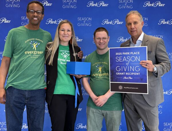 Three people in green Evergreen Life Services t-shirts with a gentleman in a gray suit representing Park Place Dealerships. He is holding a sign to announce Evergreen as a "2025 Park Place Season of Giving Grant Recipient."