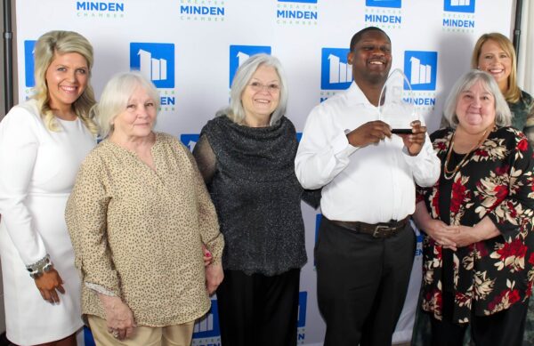 Photo of Mario holding the award along with Lessa Hamilton, Terri Howell, Carla Prioux of Evergreen and two representatives from the Greater Minden Chamber of Commerce. They are standing in front of a press wall for the Chamber of Commerce.
