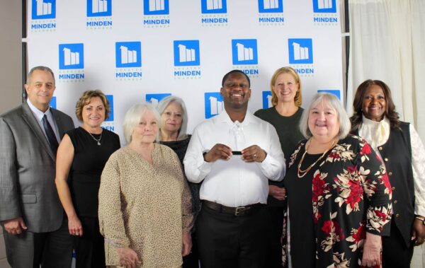 Photo of Mario holding the award along with Lessa Hamilton, Terri Howell, Carla Prioux of Evergreen and two representatives from the Greater Minden Chamber of Commerce. They are standing in front of a press wall for the Chamber of Commerce. 