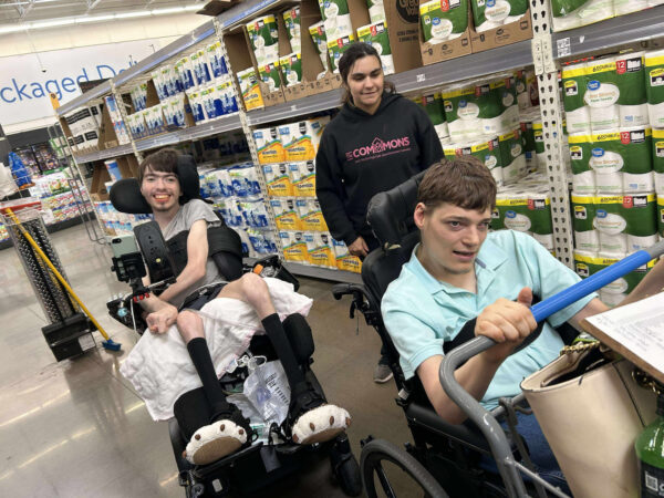 Three individuals in Oklahoma on a grocery shopping trip, two are in wheelchairs, one is standing. The person in front is pushing the cart.