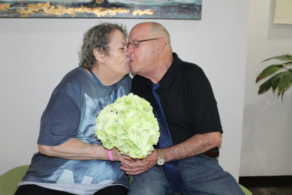 Sandra and Charles, married for 11 years, share a kiss. Sandra is seated on the left and Charles is seated on the right. They are both holding a beautiful bouquet of flowers. 
